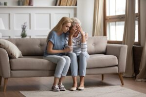 Image of two ladies sitting on a couch, hugged together. The one on the left is younger than the one on the right.