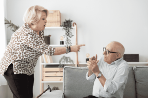 An older woman gestures towards an older man seated on a couch, highlighting a moment of interaction between them.