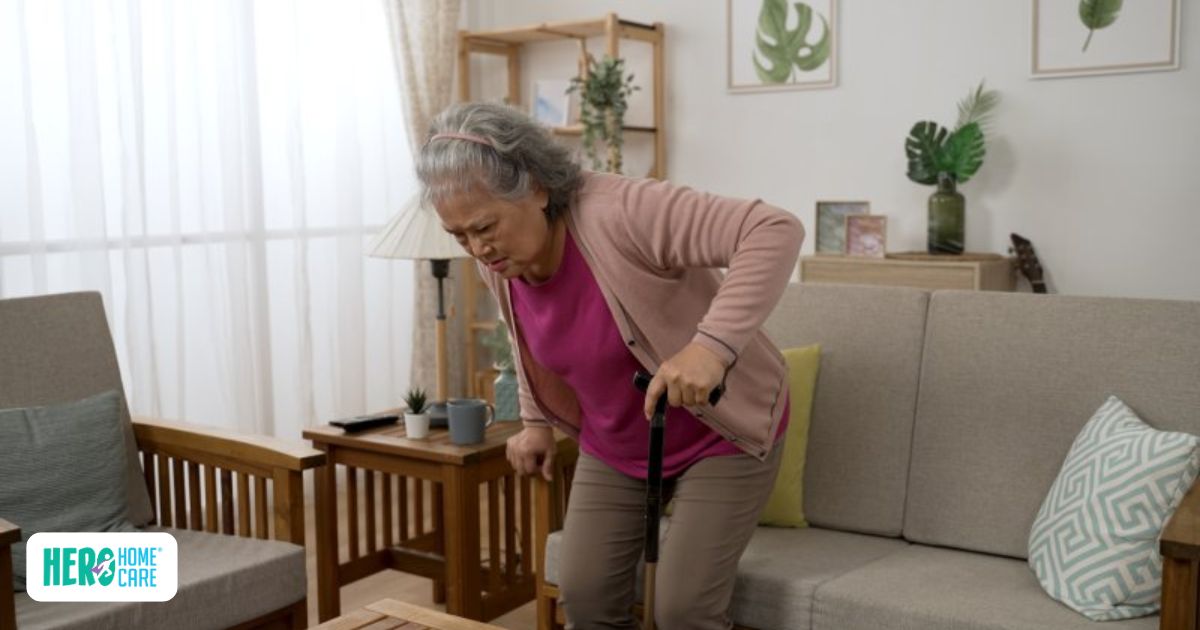 Senior woman with gray hair carefully using a cane to stand up near a living room sofa.