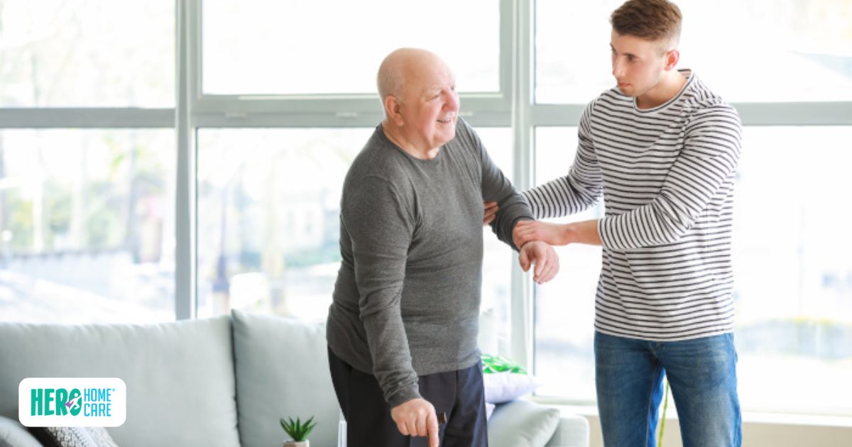 Elderly man smiling while a young caregiver gently supports his arm in a bright living room.