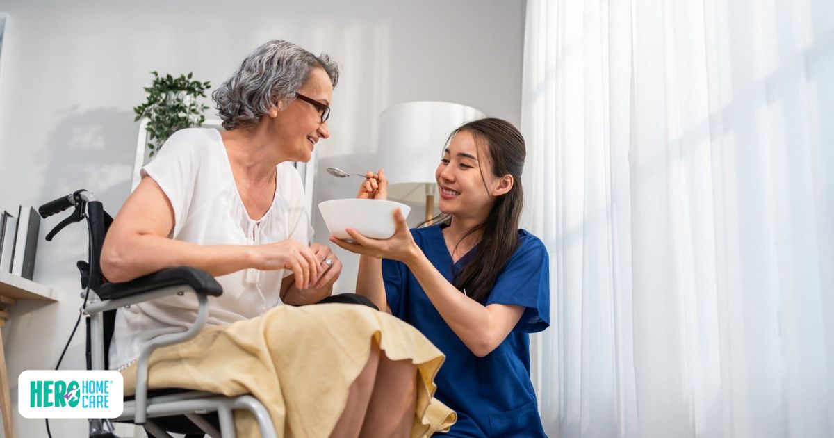 Smiling elderly woman in wheelchair being gently fed soup by a caring young caregiver in blue scrubs