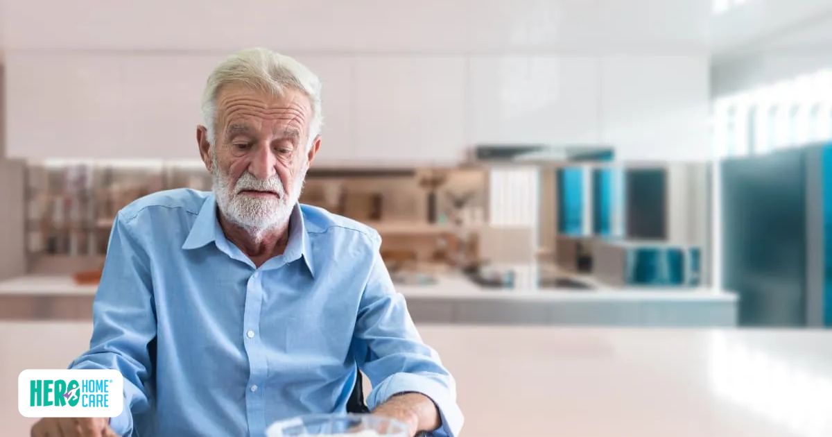 Elderly man with white beard and blue shirt sitting at a kitchen counter looking down sadly at a bowl
