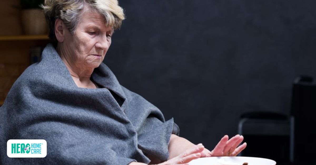 Older woman with short gray hair wrapped in a gray blanket sadly staring at an empty white plate