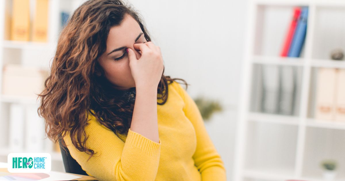 A woman sitting at a desk and rubbing the bridge of her nose with closed eyes, representing the stress and common challenges after hospital discharge like caregiver burnout.