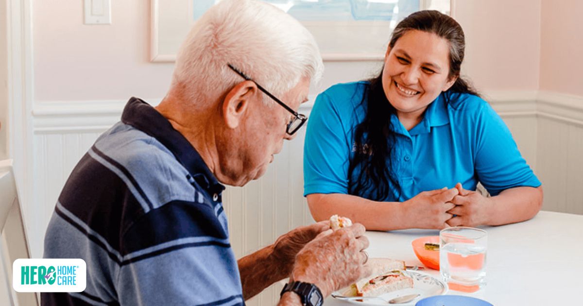 Caregiver from Hero Home Care sitting beside an elderly man at the dining table, supporting him while he eats a meal, with a warm and reassuring expression.