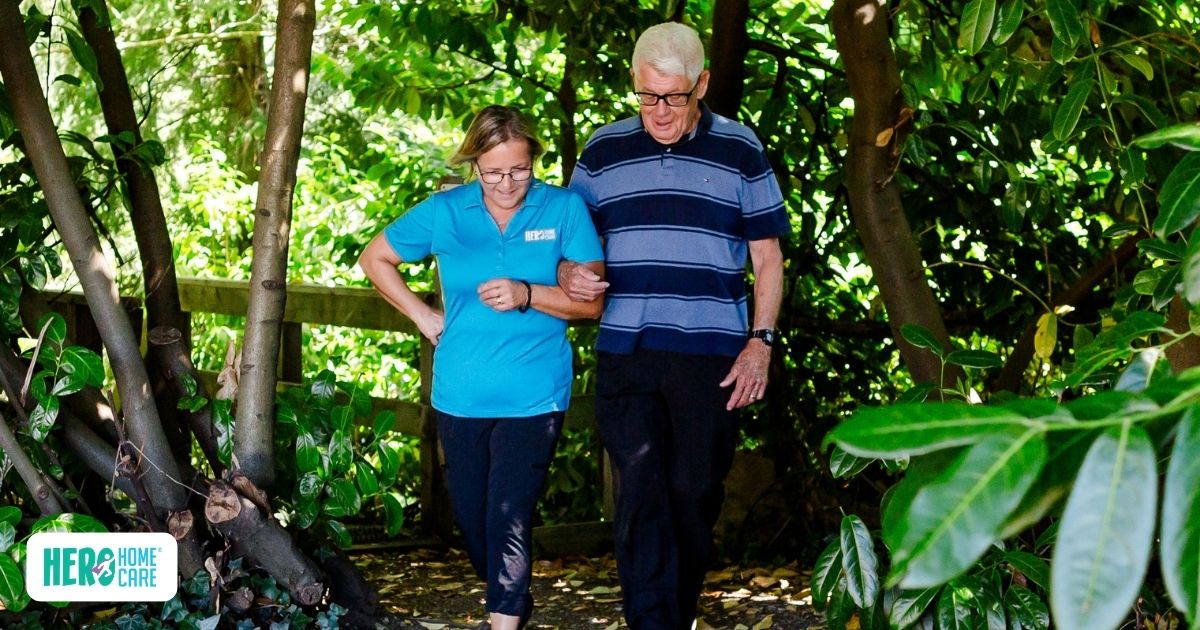Older adult walking outdoors with support from a Hero Home Care caregiver, showing mobility assistance and companionship during an outdoor activity in a green residential setting.